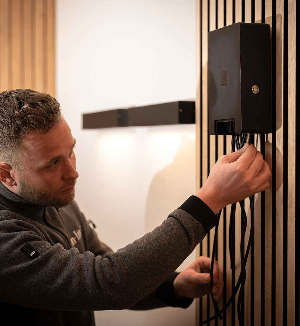 This image depicts a man adjusting or interacting with a black security device or access control system mounted on the wall. The man has a serious, focused expression as he appears to be operating or inputting something into the device. The background appears to be a dimly lit indoor space, with what looks like a wooden slatted wall or structure in the foreground. The contrast between the man's attentive posture and the subdued lighting creates a sense of purposeful, professional activity. Without being able to identify the individual, the image conveys a sense of someone engaged in a security-related task or access procedure, likely in a commercial or institutional setting. The man's body language and the presence of the security device suggest a context of monitoring, control, or entry management. Overall, the image captures a moment of a person interacting with a security system, though the specifics of the situation or location are not clear from the visual information provided.