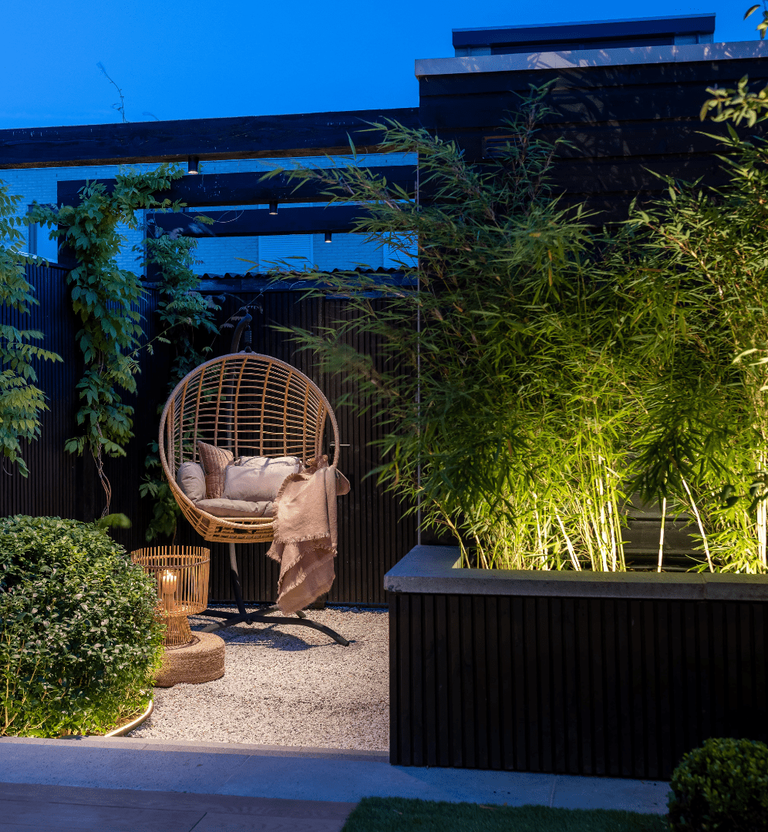 This image depicts a beautifully designed and curated outdoor living space, featuring a harmonious blend of contemporary architectural elements and lush, natural vegetation.  The focal point is a cozy, woven rattan egg chair positioned on a raised platform or deck. The chair is adorned with plush, inviting cushions, creating a comfortable and visually appealing seating area. Surrounding the chair, the space is filled with verdant, thriving plants and bamboo shoots, creating a sense of privacy and immersion in the natural environment.  The architectural elements, including the black wooden frame and overhead structures, provide a sleek, modern contrast to the organic, living components. The combination of the dark, clean lines and the vibrant, textured foliage results in a visually striking and harmonious composition.  The overall design emphasizes the integration of the designed and natural elements, creating a serene and inviting outdoor oasis that blends seamlessly with the surrounding landscape. The use of natural materials, such as the rattan and wooden structures, further enhances the connection between the man-made and the natural.  This image conveys a sense of elegance, comfort, and a deep appreciation for the beauty of the outdoors, where the built environment and the living landscape coexist in a cohesive and aesthetically pleasing manner.