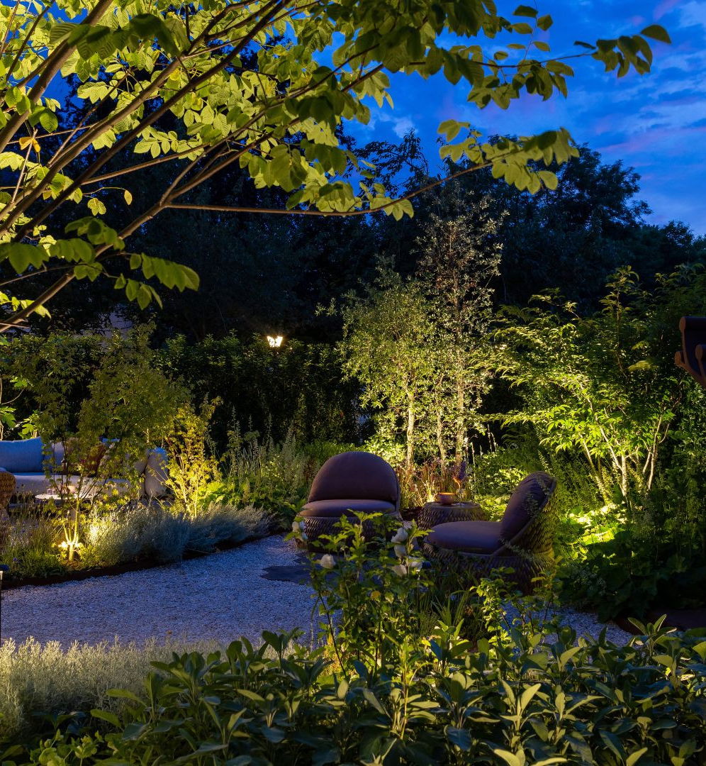 This image depicts a lush and tranquil outdoor garden scene at night. The focal point is a cozy seating area surrounded by a variety of vibrant, well-maintained plants and trees. The seating area features comfortable-looking cushioned chairs and a low table, creating an inviting space for relaxation and entertainment. The entire garden is illuminated by strategically placed landscape lighting, which casts a warm, ambient glow over the scene and highlights the diverse foliage and textures of the plants. The clear, starry night sky in the background adds to the serene and peaceful atmosphere of the space. Overall, this image conveys a sense of harmonious integration between the designed elements and the natural environment, resulting in a visually captivating and inviting outdoor oasis.